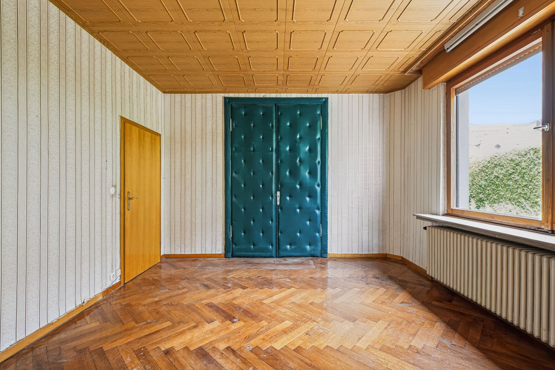 Leerer Raum mit Parkettboden, holzvertäfelter Decke, grün gepolsterter Tür und großem Fenster.
