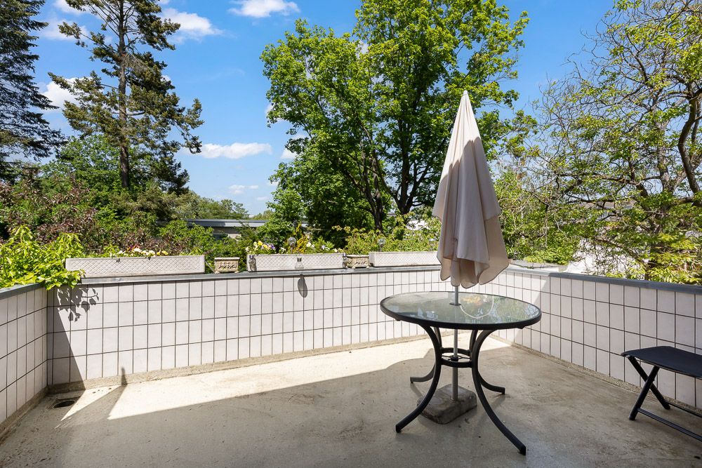 Balkon mit rundem Glastisch, geschlossenem Sonnenschirm und Blick auf grüne Bäume.