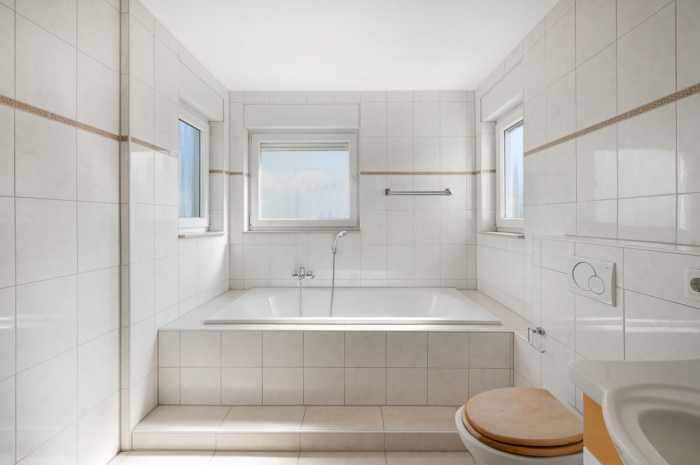 Helles Badezimmer mit Badewanne, WC, Waschbecken und drei Fenstern.