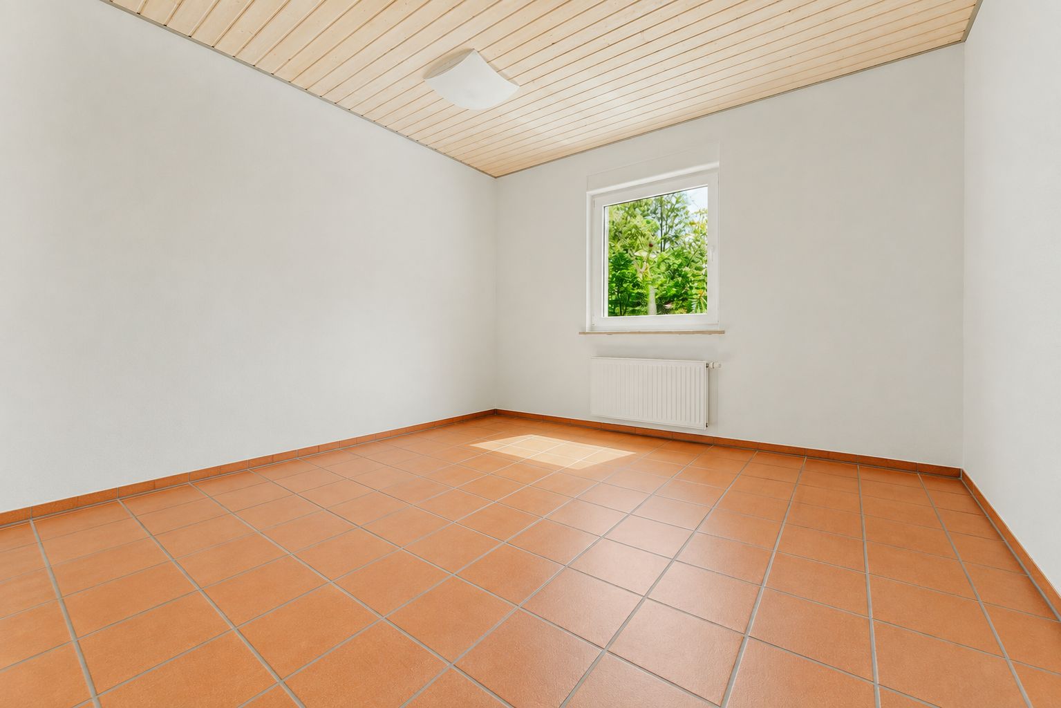 Leeres Zimmer mit weißen Wänden, Fliesenboden, Holzdecke und Fenster mit Blick ins Grüne.