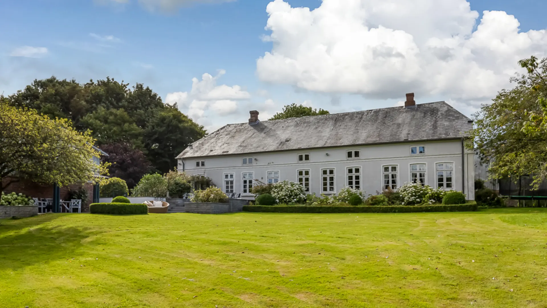 Weißes Wohnhaus mit großen Fenstern, Garten und Rasenfläche, umgeben von Bäumen.
