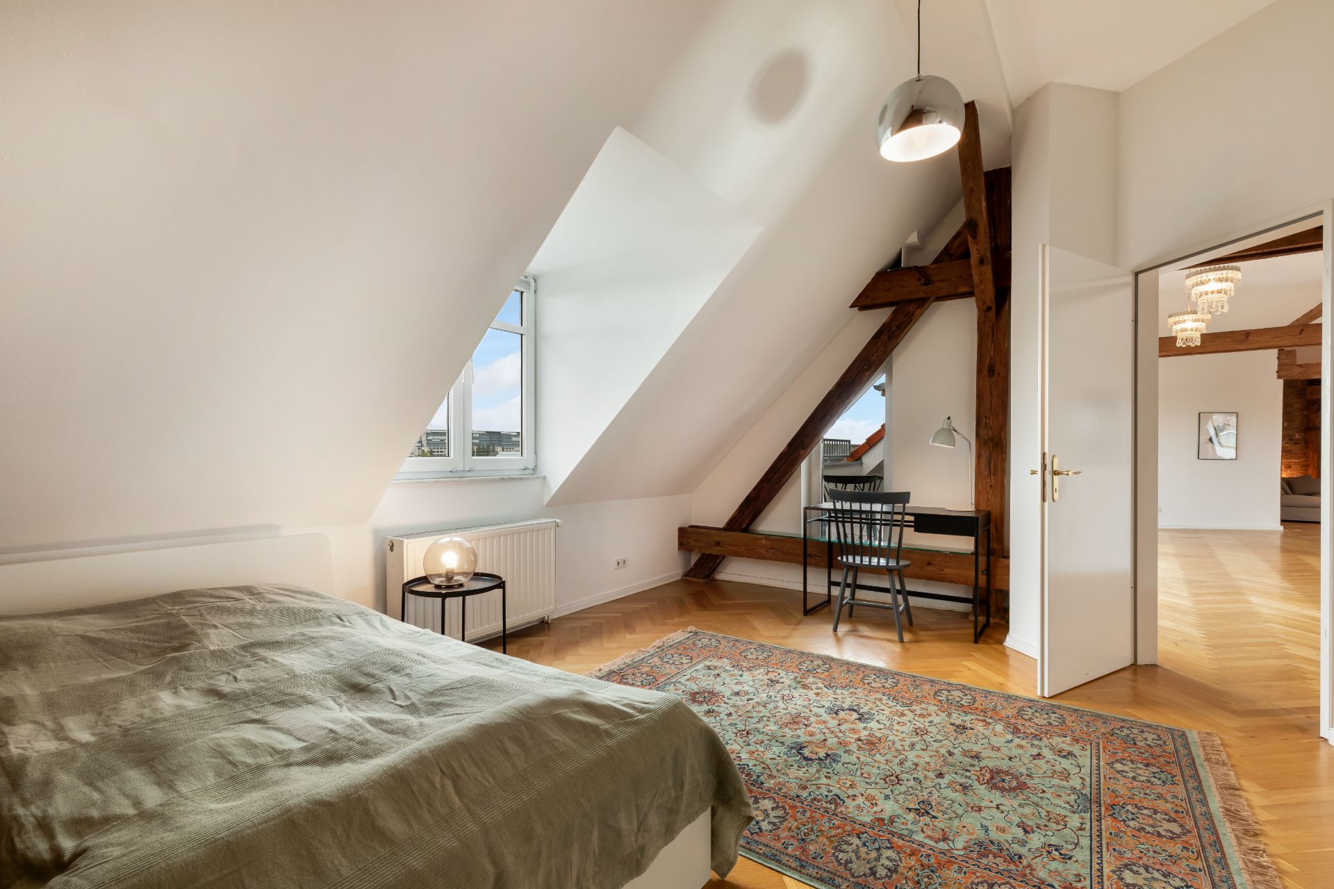 Schlafzimmer mit Teppich, Bett, Schreibtisch und großem Fenster unter Dachschrägen.