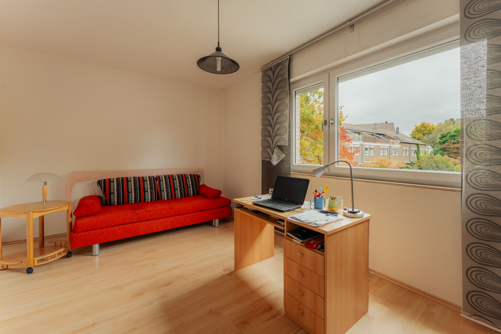 Helles Zimmer mit rotem Sofa, Schreibtisch und großem Fenster mit Blick nach draußen.