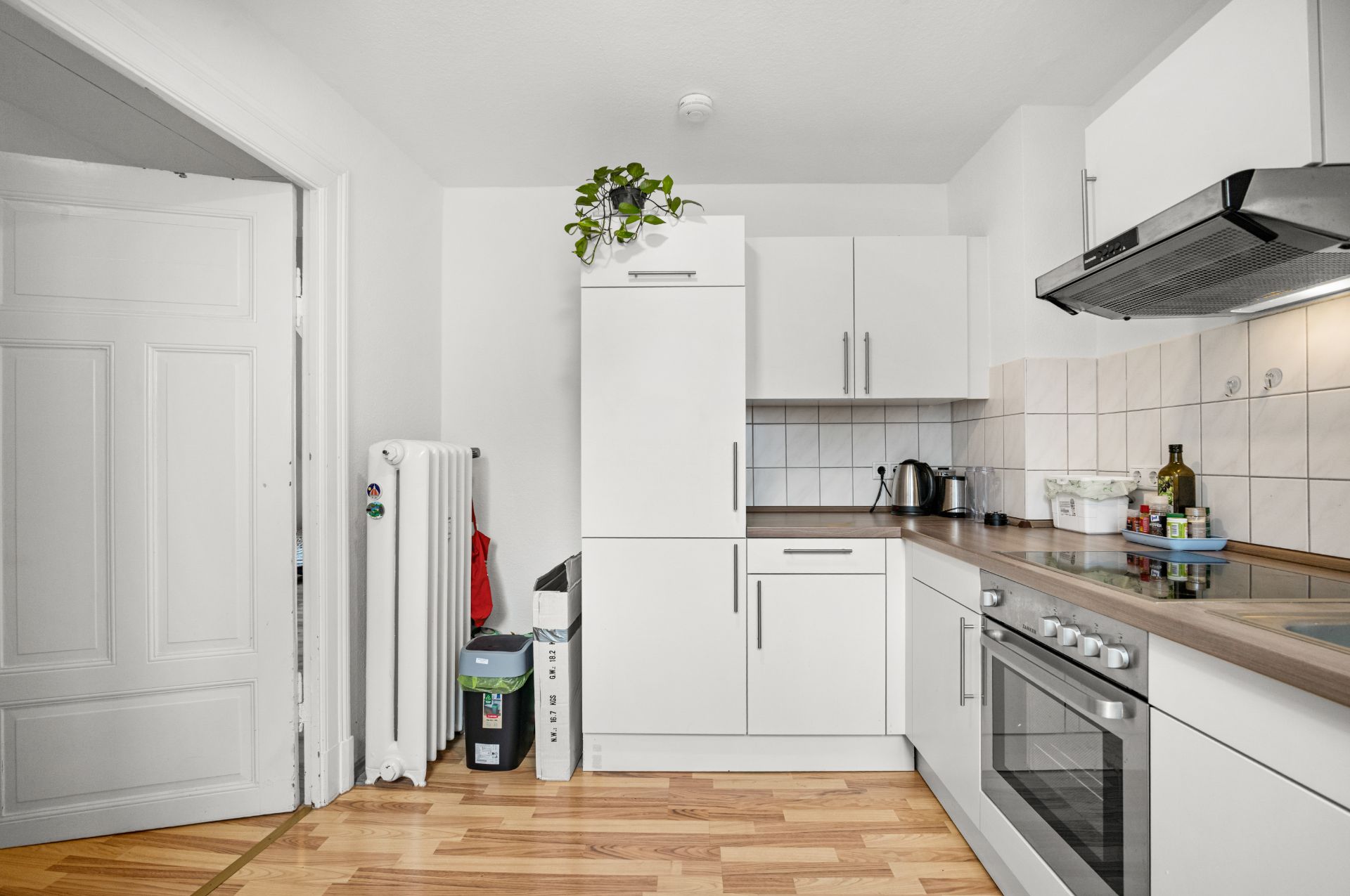 Moderne weiße Einbauküche mit Holzfußboden, Elektrogeräten und offenem Türrahmen.