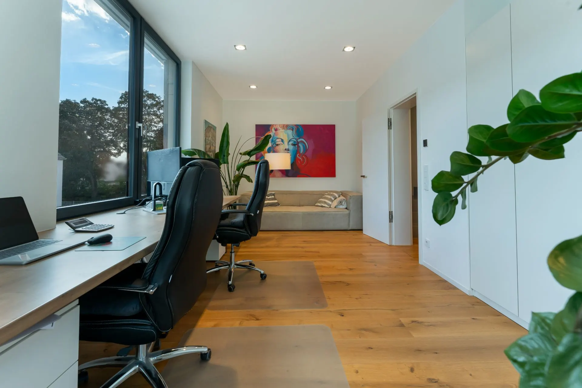 Modernes Arbeitszimmer mit großem Fenster, Schreibtisch, zwei Stühlen, Sofa und Pflanzen.