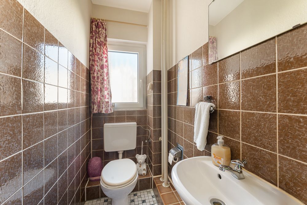 Kleines Badezimmer mit braunen Fliesen, WC, Waschbecken und Fenster mit geblümtem Vorhang.