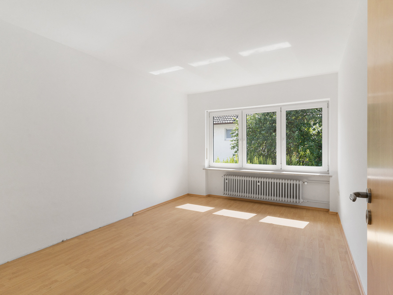 Heller leerer Raum mit Holzboden, weißer Wand und großem Fenster mit Blick ins Grüne.