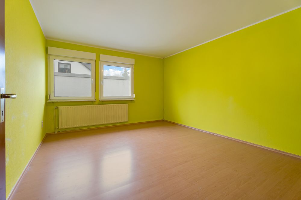 Leeres Zimmer mit gelben Wänden, zwei Fenstern und hellem Laminatboden.