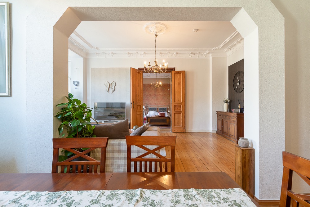 Esszimmer mit Holztisch, Blick ins Wohnzimmer mit Sofa, Pflanzen und Kronleuchter.