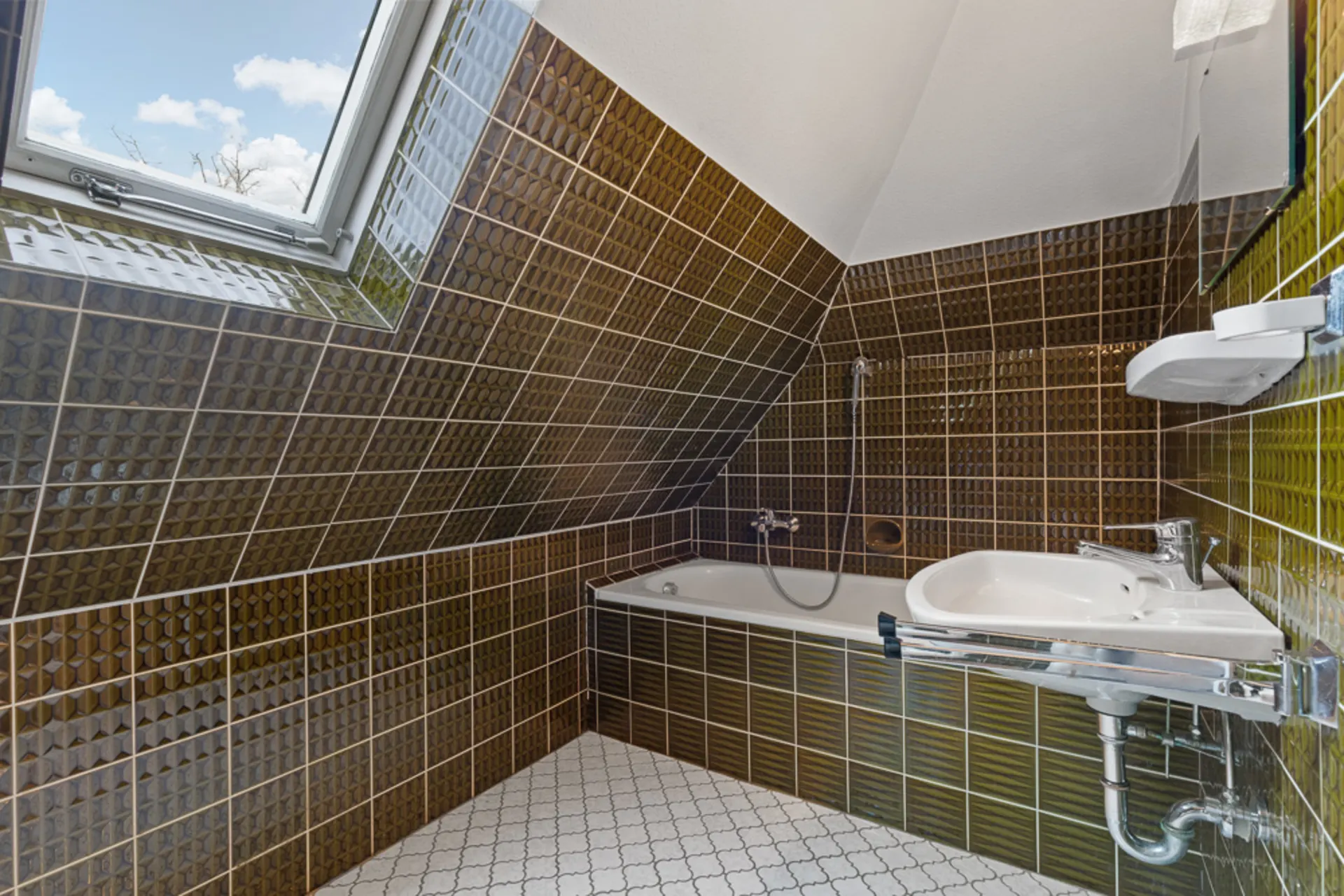 Badezimmer mit braunen Fliesen, Badewanne, Waschbecken und Fenster in der Dachschräge.