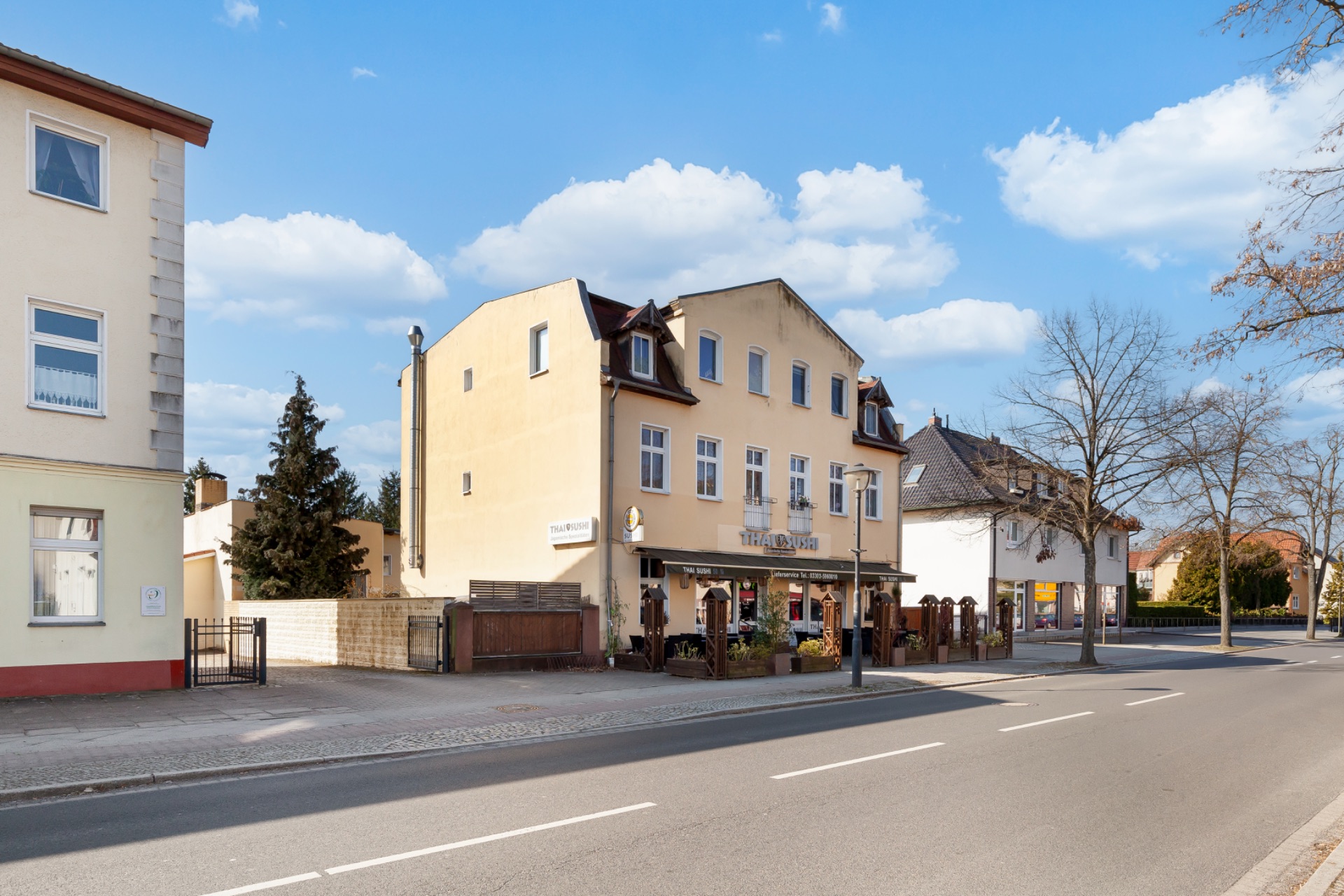 Zweistöckiges Wohnhaus mit Terrasse an einer ruhigen Straße, Bäume und Gehweg im Vordergrund.
