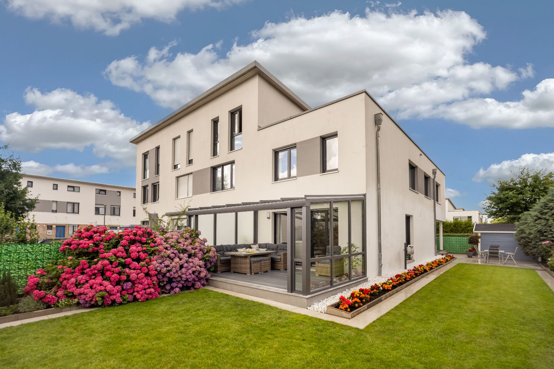Modernes Wohnhaus mit Wintergarten, gepflegtem Garten, Blumenbeeten und Sitzbereich im Freien.