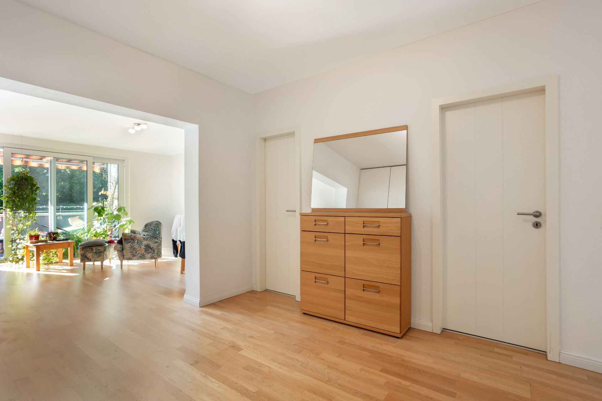 Flur mit Holzkommode und Spiegel, zwei Türen und Blick ins helle Wohnzimmer mit Pflanzen.