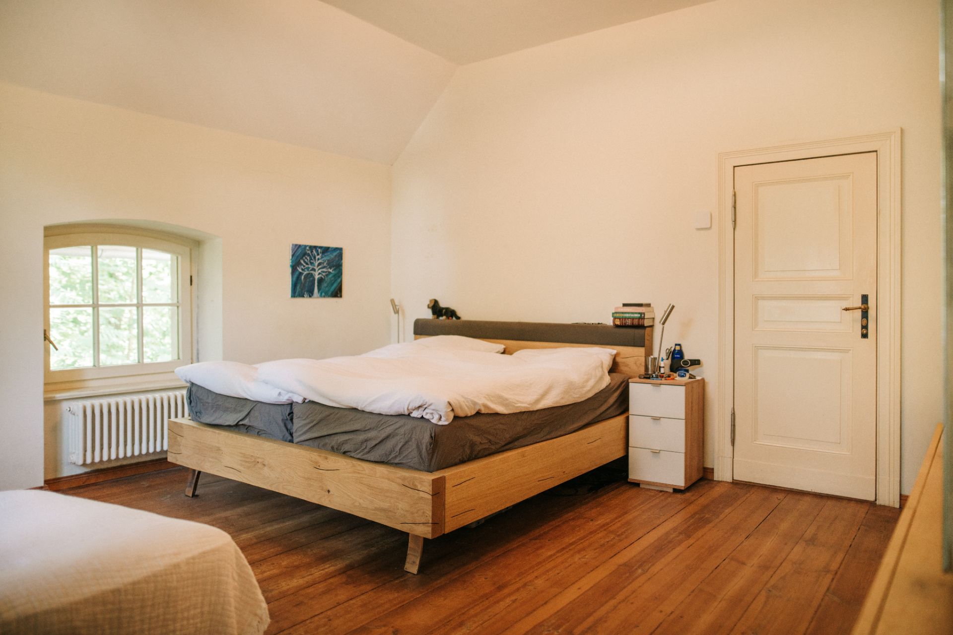 Schlafzimmer mit Doppelbett, Nachttisch, Holzboden, Fenster und weißer Tür.
