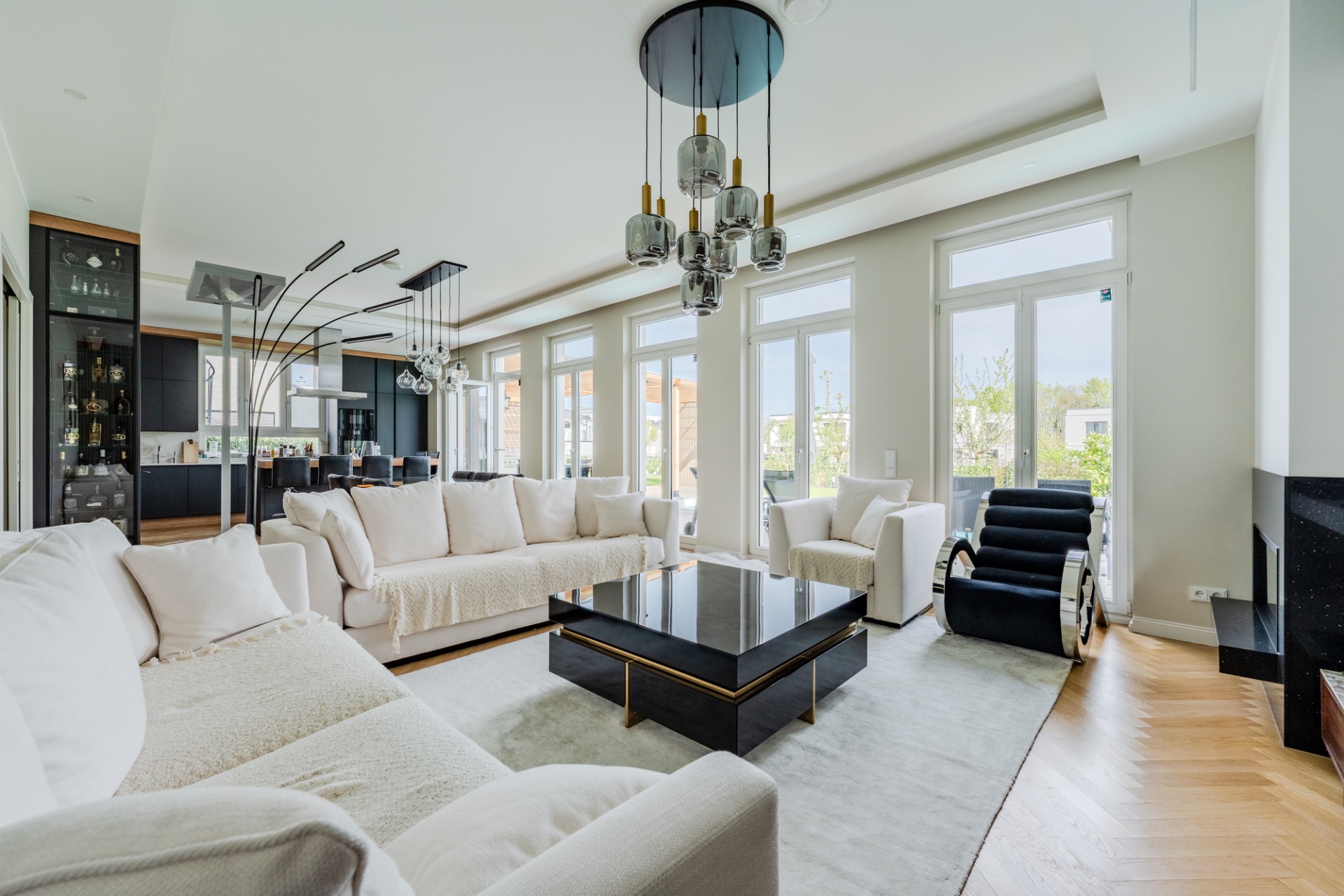 Modernes Wohnzimmer mit weißen Sofas, großem Couchtisch und großen Fenstern, Blick auf Essbereich und Küche.