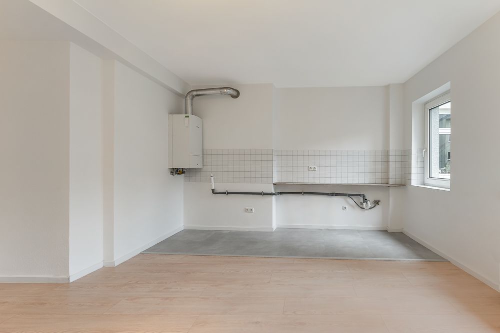 Leere Küchenzeile mit weißen Fliesen, Gastherme, Anschlüssen und Fenster auf der rechten Seite.