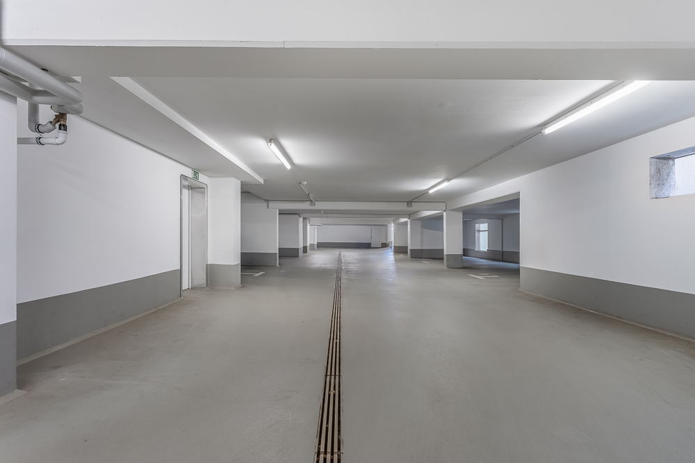 Leerer, heller Tiefgaragenraum mit weißen Wänden und grauem Boden.