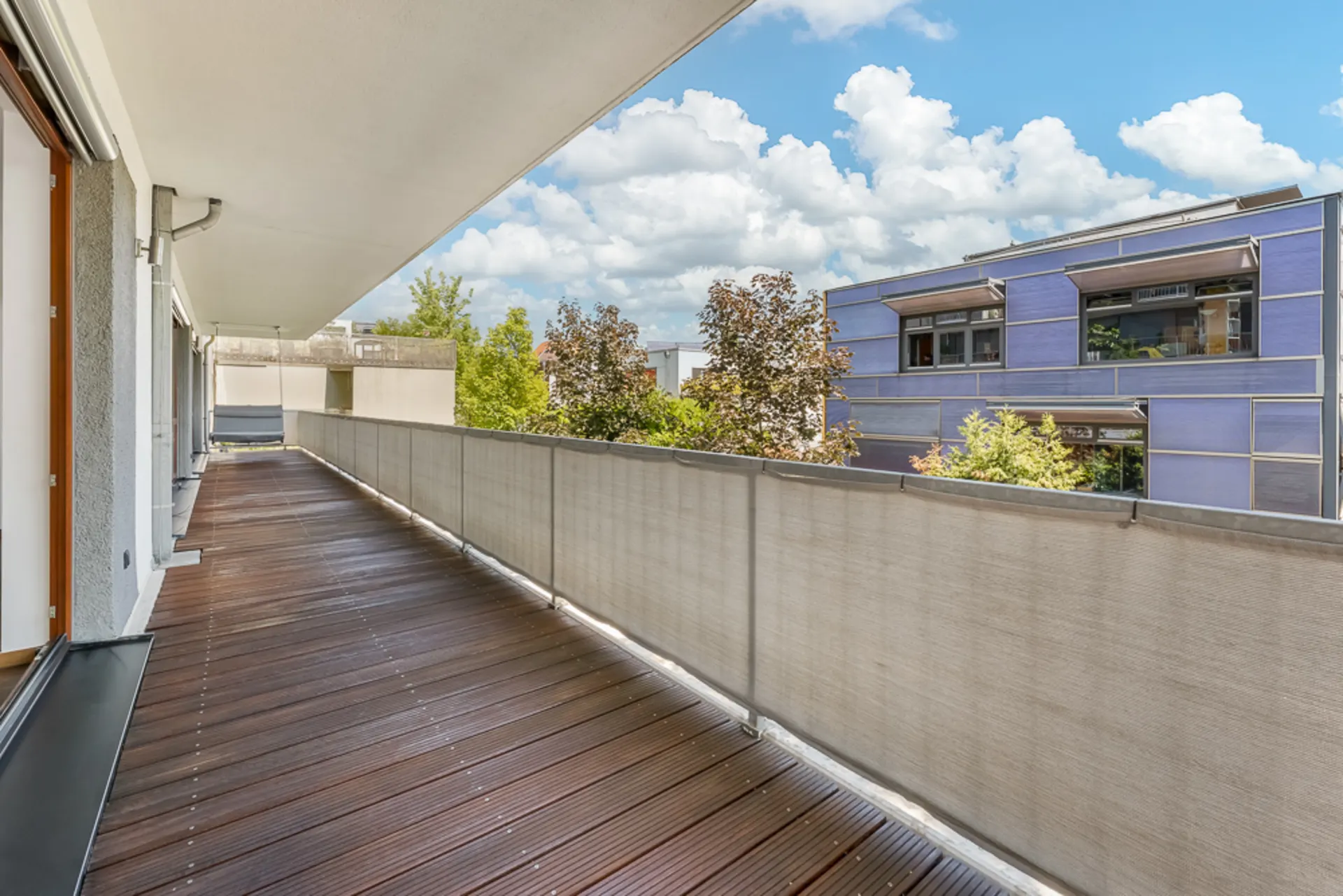 Großer Balkon mit Holzfußboden und Blick auf ein modernes Wohngebäude und Bäume.
