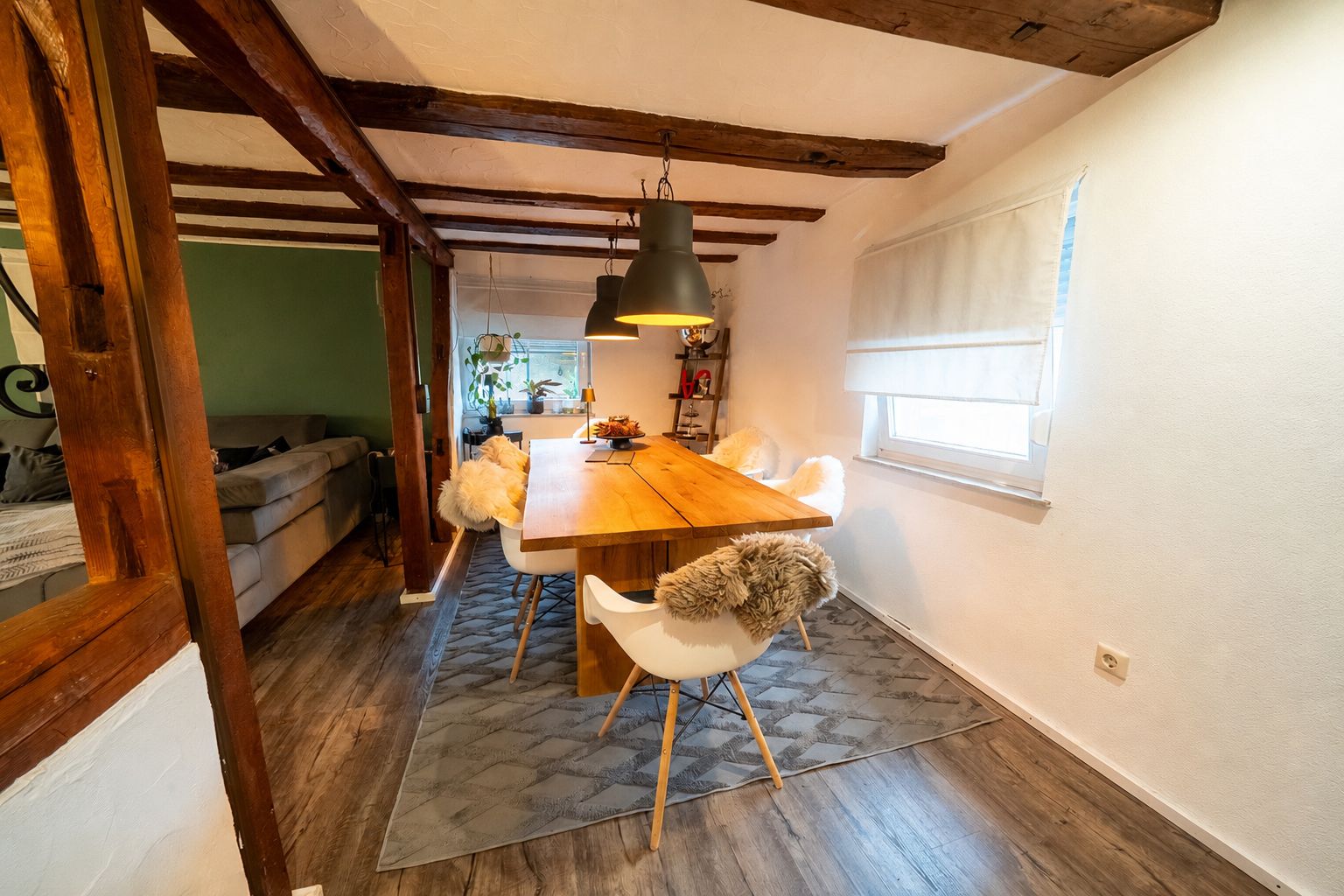 Essbereich mit Holztisch, weißen Stühlen und Teppich, sichtbare Holzdeckenbalken.