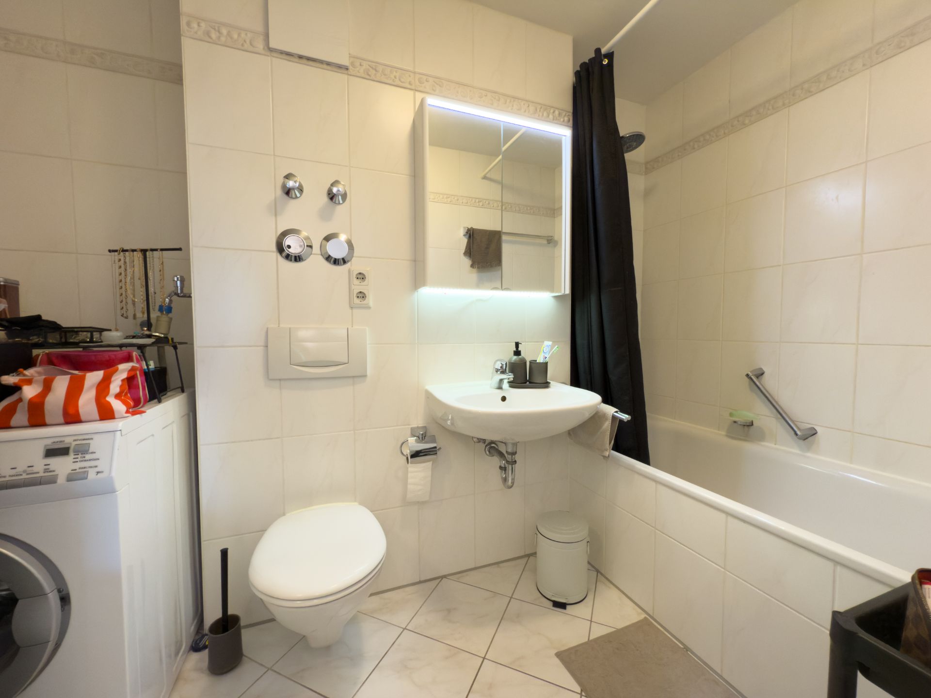 Helles Badezimmer mit Waschmaschine, Toilette, Waschbecken, Spiegel und Badewanne mit Duschvorhang.