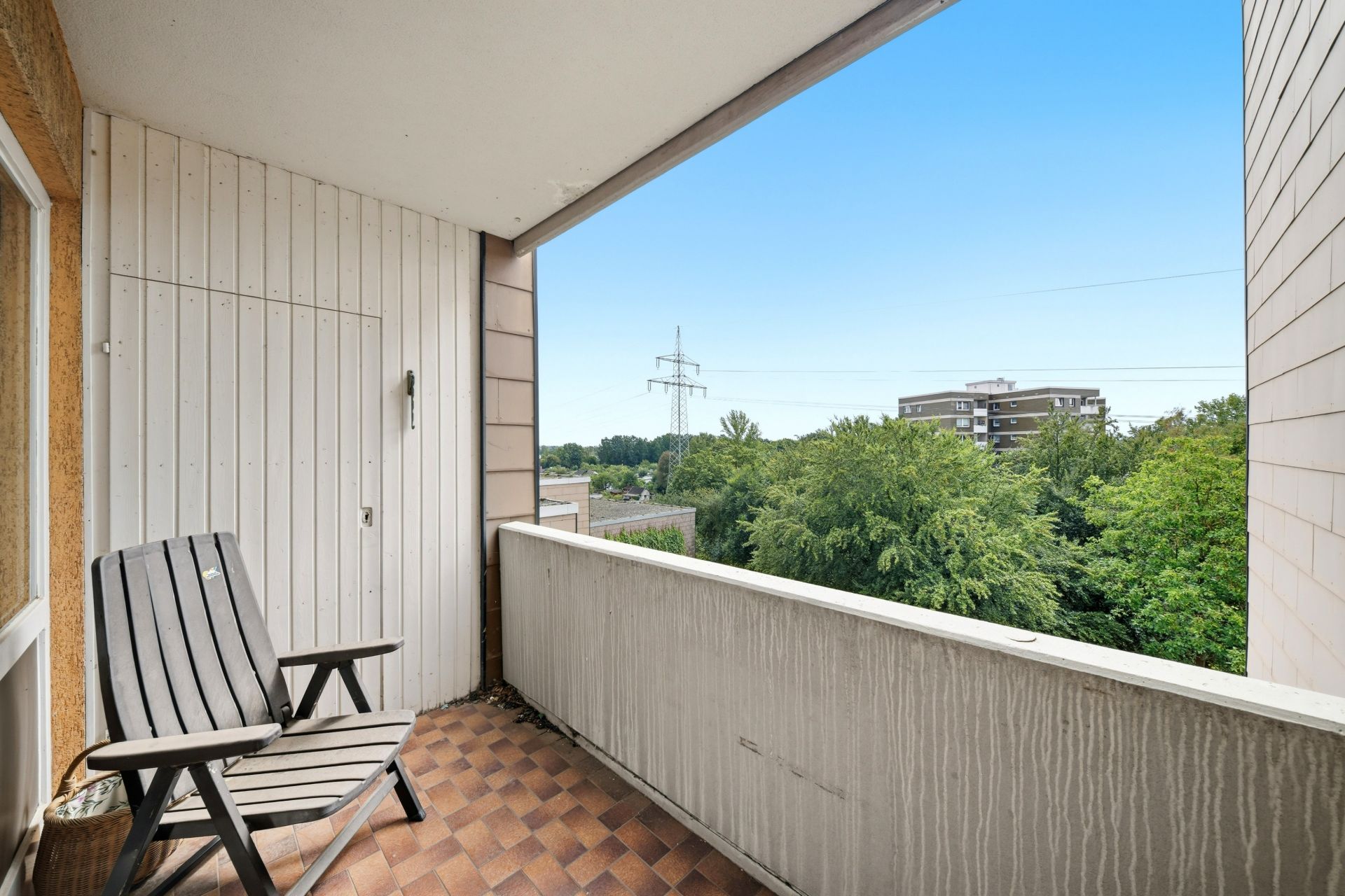 Balkon mit Holzwand, Stuhl und Blick ins Grüne sowie auf Wohngebäude.