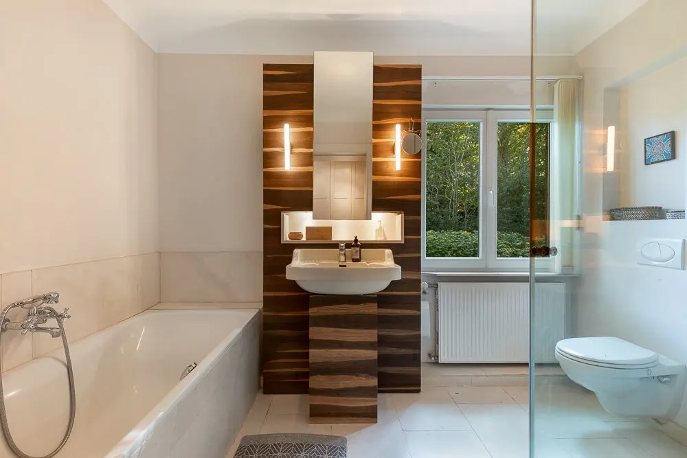 Modernes Badezimmer mit Badewanne, Waschbecken, Wandspiegel, Fenster und Wandtoilette.