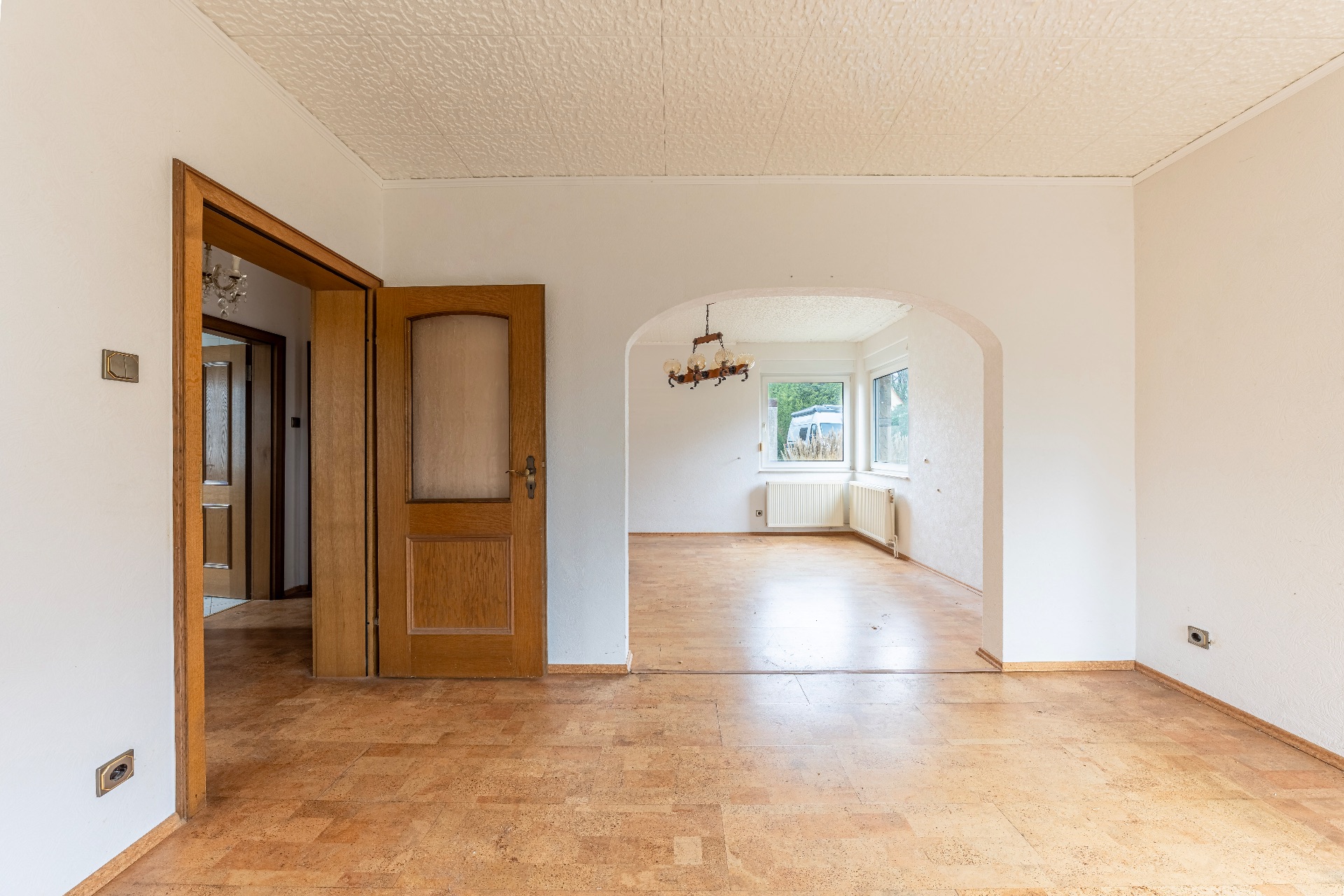 Leerer Wohnraum mit Holztür, offener Durchgang und großem Fenster im Hintergrund.