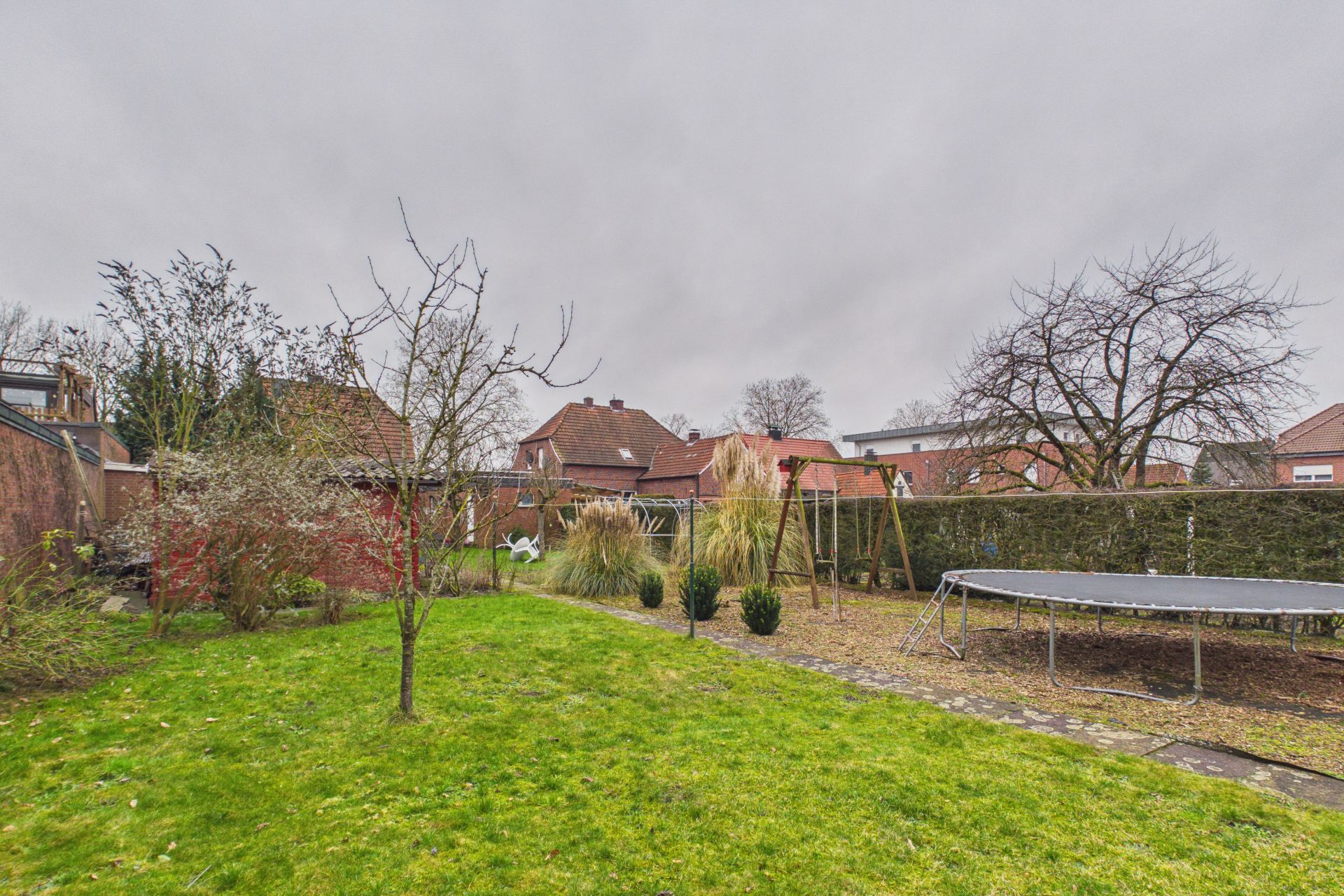 Garten mit Rasen, Bäumen, Sträuchern, Schaukel und Trampolin unter grauem Himmel.