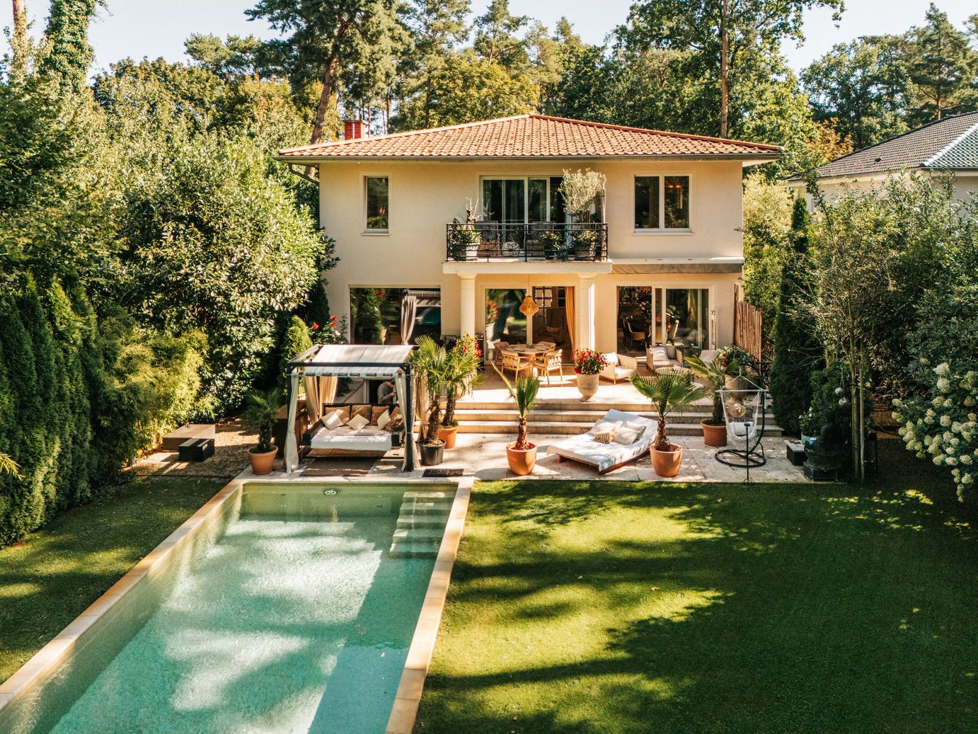Zweistöckiges Haus mit Garten, Terrasse, Pool und Sonnenliegen, umgeben von Bäumen.