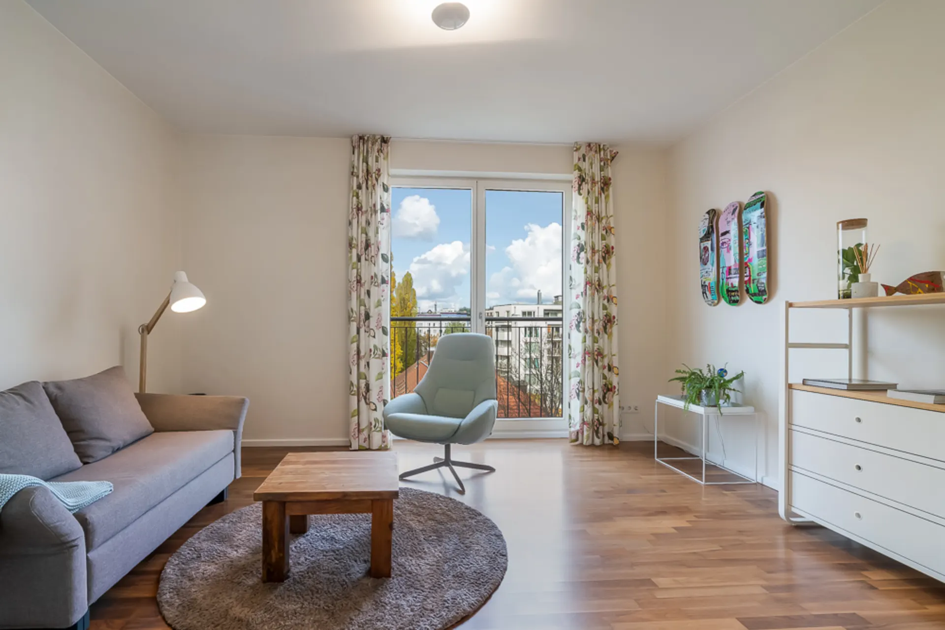 Helles Wohnzimmer mit Sofa, Sessel, Couchtisch und großem Fenster mit Vorhängen.