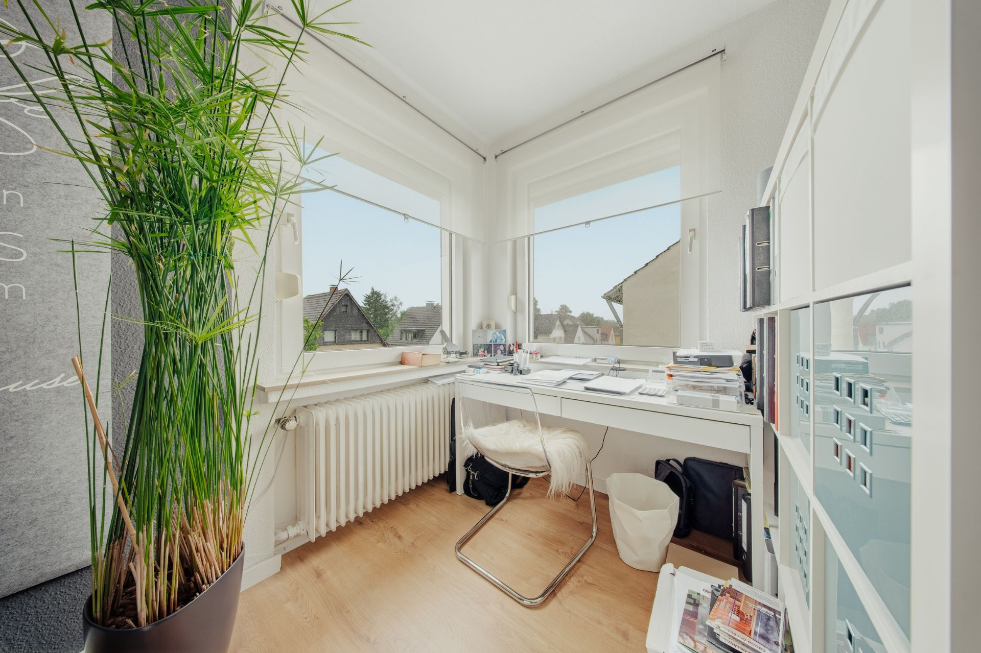 Helles Arbeitszimmer mit Eckschreibtisch, Stuhl, großen Fenstern und einer hohen Pflanze.