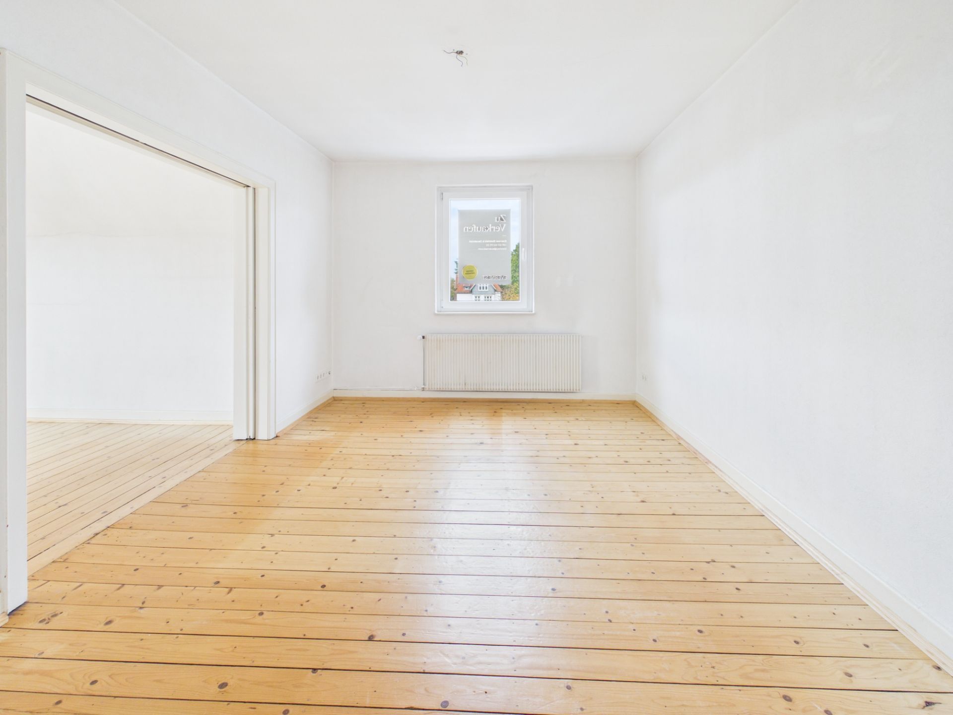 Leerer Raum mit Holzboden, weiß gestrichenen Wänden und einem Fenster mit Heizkörper.