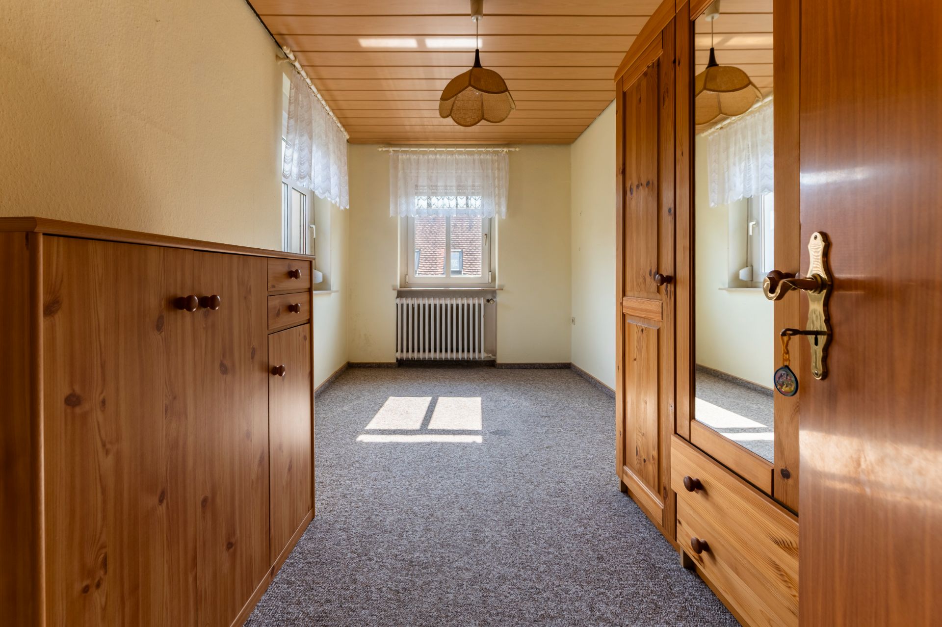 Schmaler Raum mit Holzmöbeln, zwei Fenstern, Teppichboden und Deckenlampe.