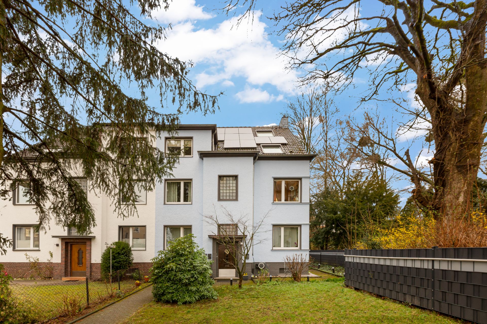 Mehrfamilienhaus mit weißer Fassade, mehreren Fenstern und Vorgarten mit Bäumen und Sträuchern.