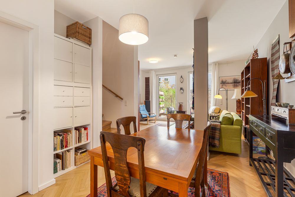 Essbereich mit Holztisch, Bücherregal, grünem Sofa und Zugang zum Balkon in einem Wohnraum.