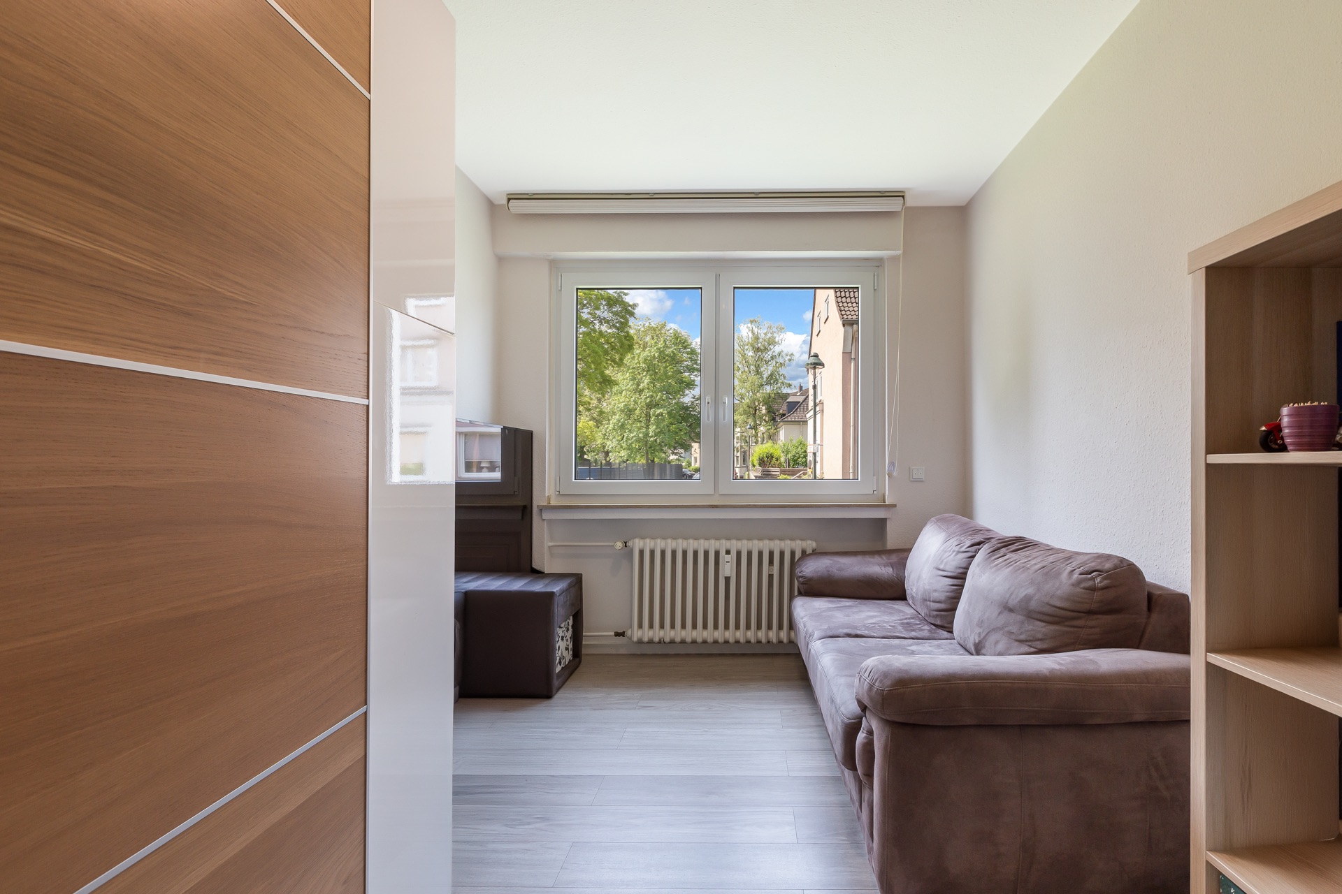 Wohnzimmer mit braunem Sofa, Regal und großem Fenster mit Blick ins Grüne.