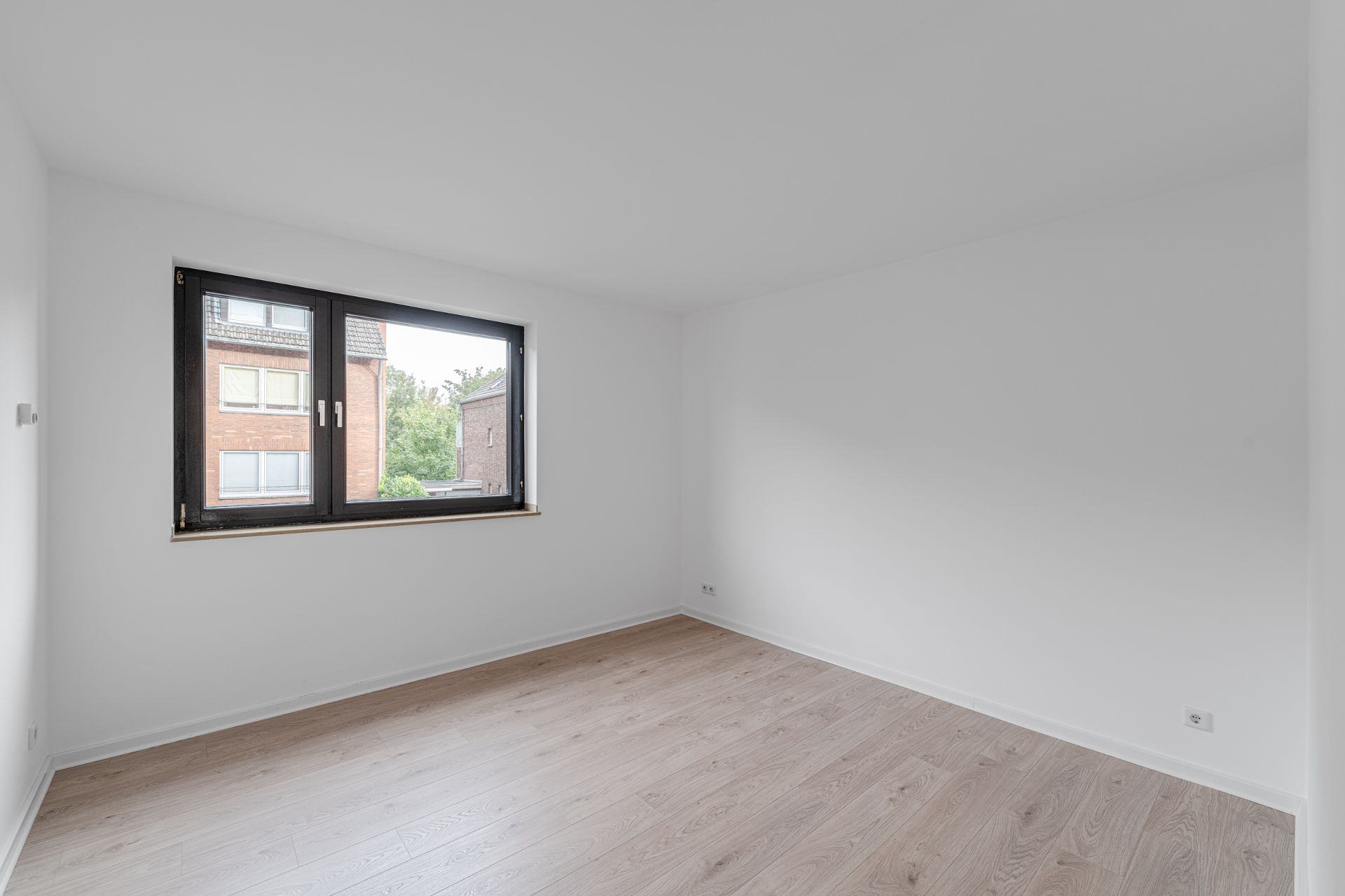 Leerer Raum mit hellem Holzfußboden und großem Fenster, weiße Wände.