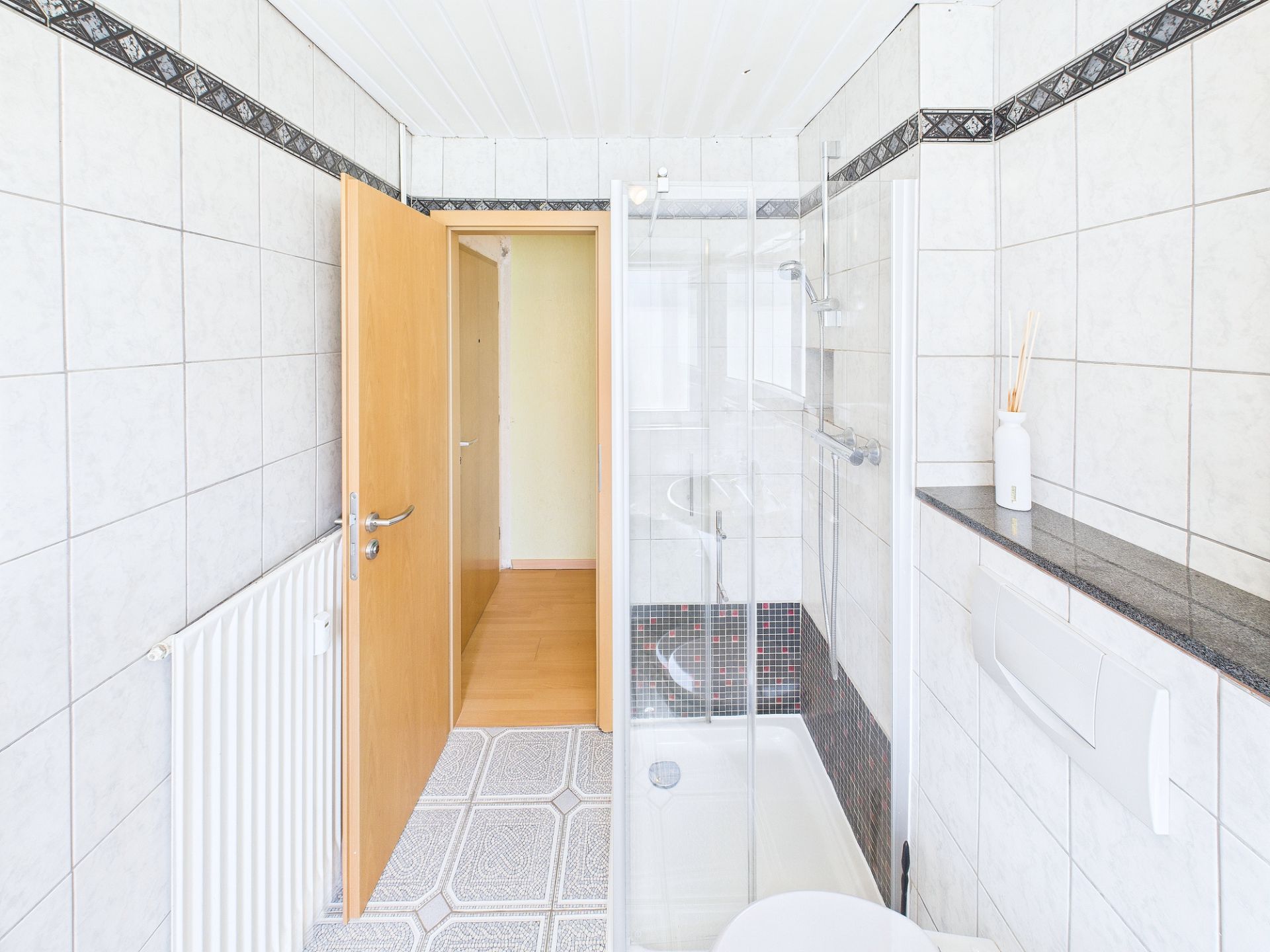 Helles Badezimmer mit Dusche, weißen Fliesen und offen stehender Holztür.