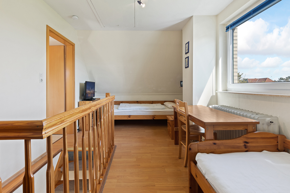 Helles Zimmer mit Holztreppe, Bett, Tisch, Stühlen und großem Fenster.