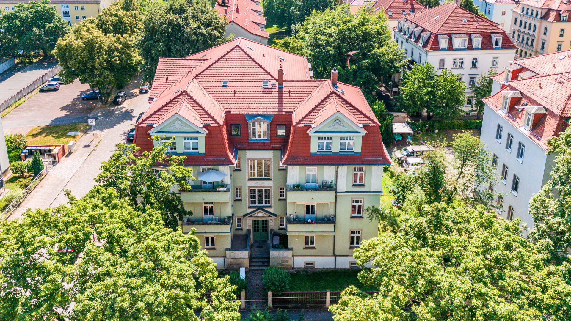 Mehrfamilienhaus mit rotem Dach, Balkonen und von Bäumen umgeben, aus der Vogelperspektive.