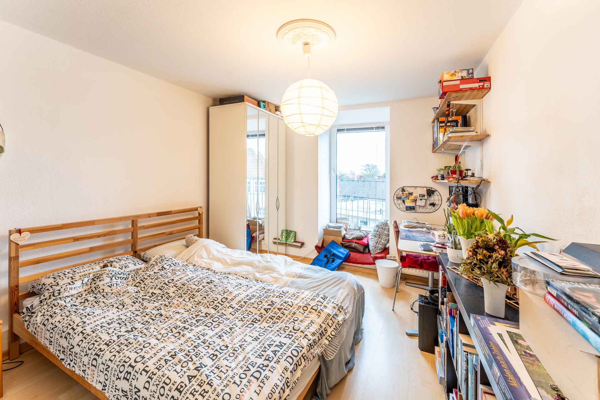 Helles Schlafzimmer mit Doppelbett, Schreibtisch, Regal und großen Fenstern.