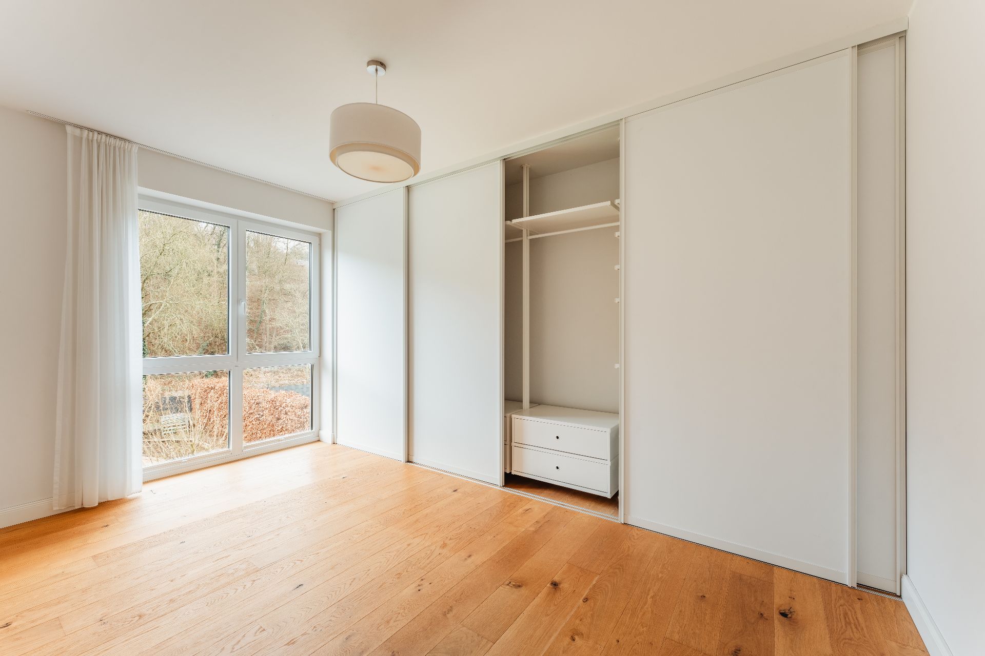 Helles, leeres Zimmer mit Holzfußboden, großem Fenster und Einbauschrank.