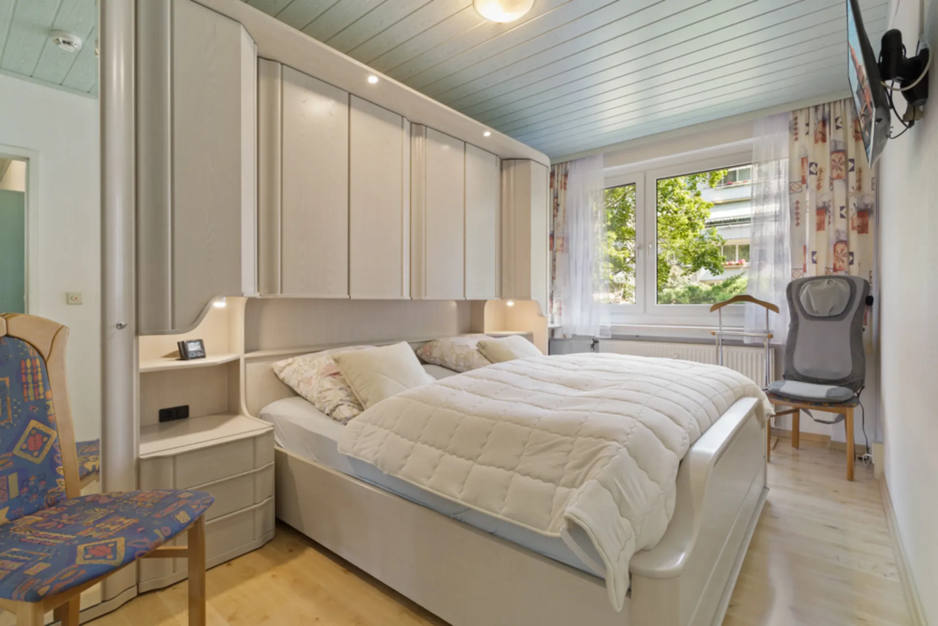 Helles Schlafzimmer mit Doppelbett, Einbauschränken und großem Fenster mit Vorhängen.