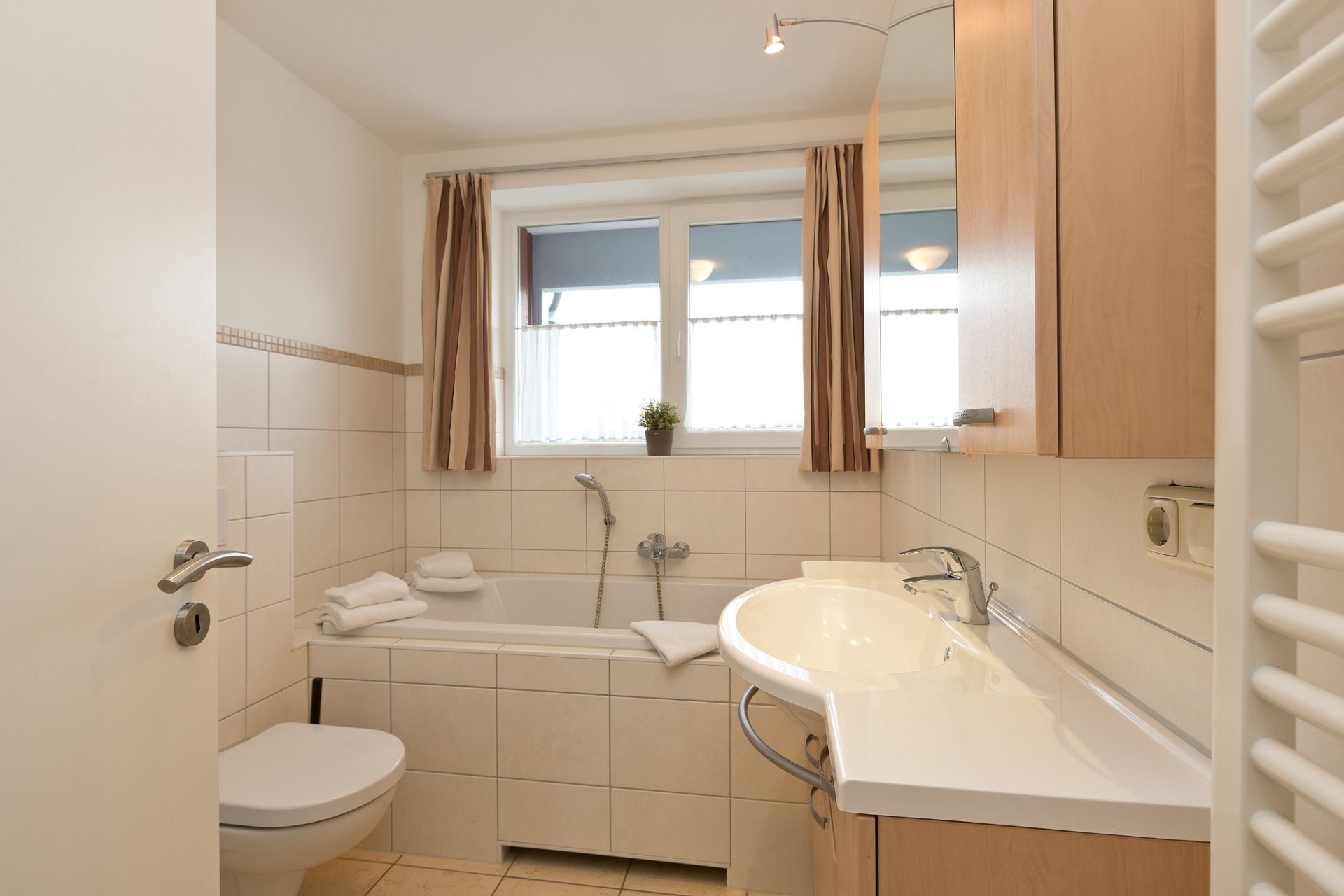 Helles Badezimmer mit Badewanne, Waschbecken, WC und Fenster mit Vorhängen.