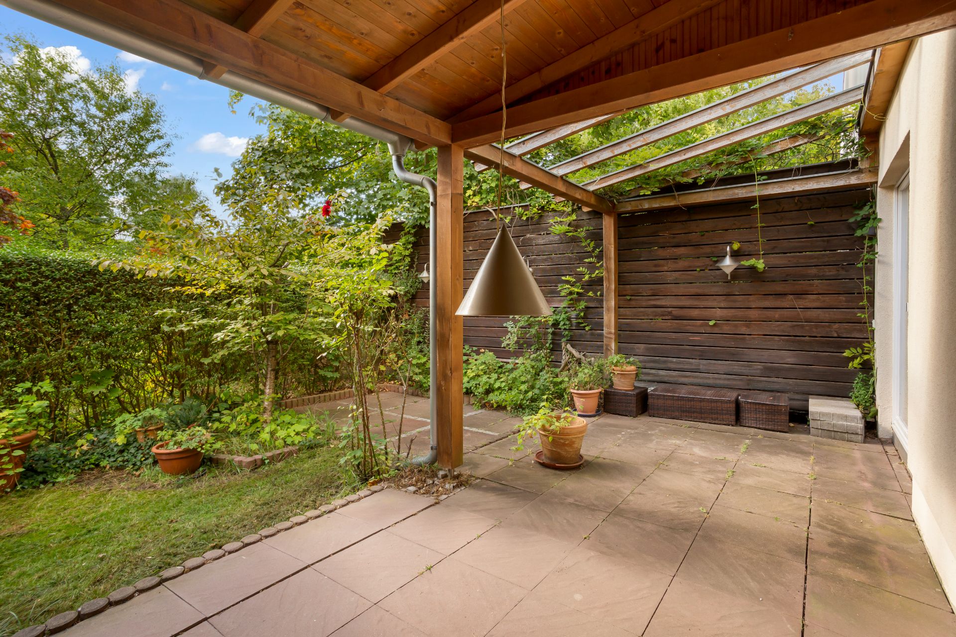 Überdachte Terrasse mit Holzdach, Pflanzen in Töpfen und Blick auf einen Garten mit Hecke.