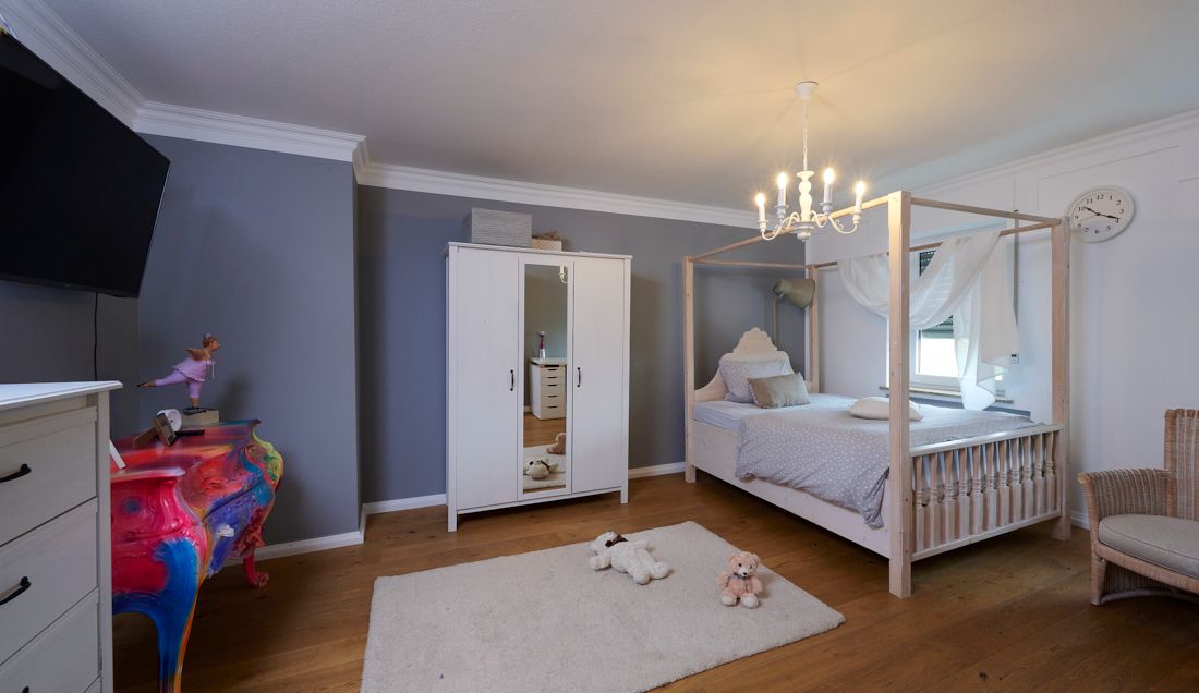 Kinderzimmer mit Himmelbett, Kleiderschrank, bunter Kommode und Plüschtieren auf Teppich.