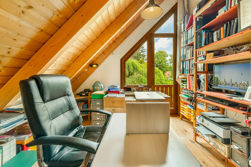 Helles Arbeitszimmer mit Schreibtisch, Bürostuhl, Regalen und Dachschrägen, großes Fenster mit Gartenblick.