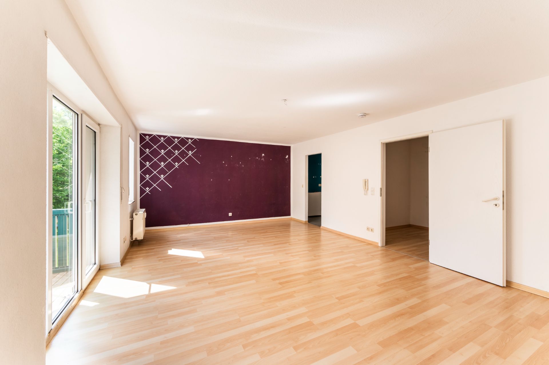 Helles, leeres Wohnzimmer mit Holzfußboden, großer Fensterfront und lila Akzentwand.