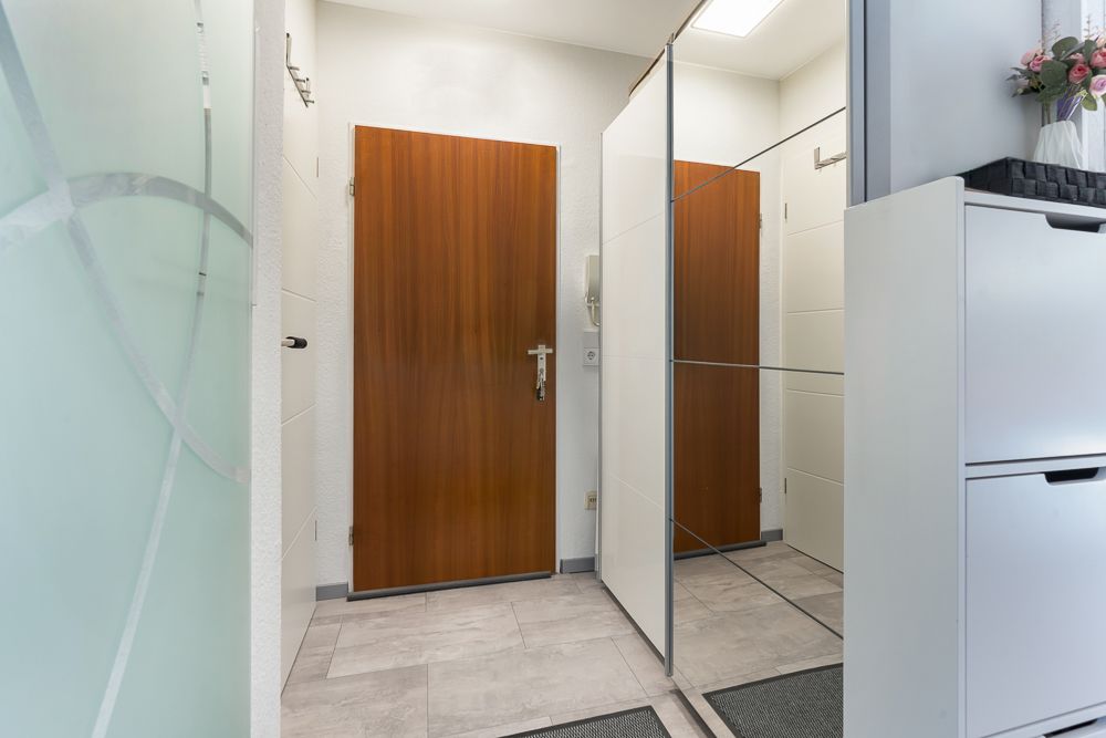 Eingangsbereich mit Holztür, Spiegelschrank, weißem Schuhschrank und Fliesenboden.