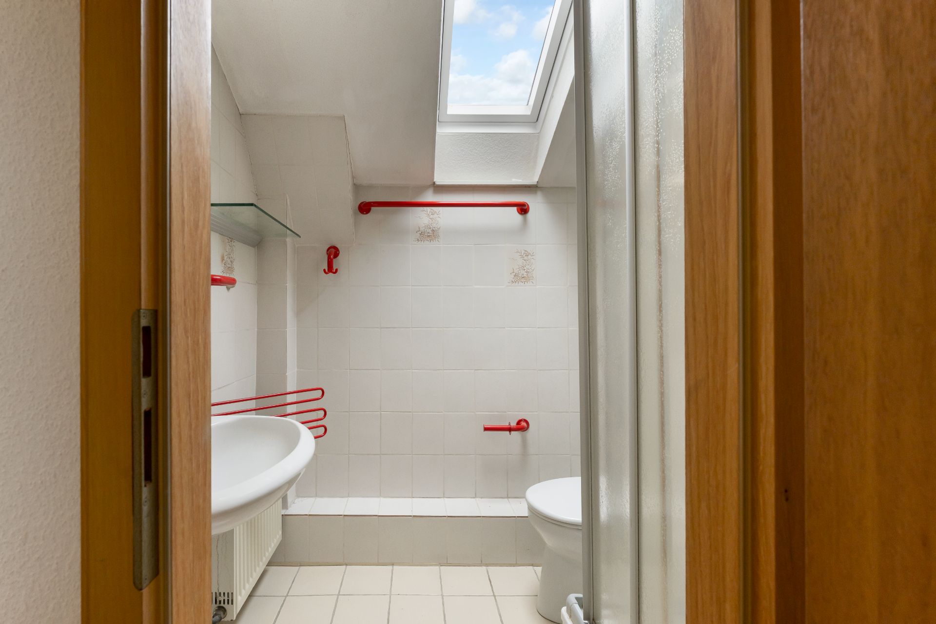 Kleines Badezimmer mit Dachfenster, rotem Haltegriff, Waschbecken und Toilette.